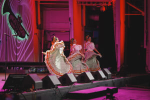 Three women in colorful traditional dresses perform a folkloric dance on a pink-lit stage with microphones nearby.