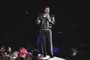 Male performer in a black, gold-embroidered jacket sings into a handheld mic on a dark stage, with front-row audience visible.