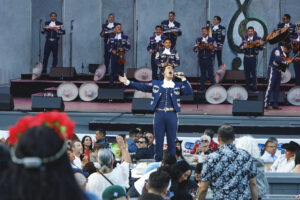 Male mariachi singer in a blue suit performs center stage with a brass-and-string ensemble behind, arms raised to the crowd.