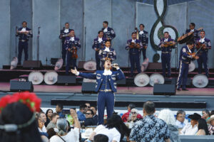 Male mariachi singer center stage with a full blue-outfit band performing to a seated crowd at an outdoor show.