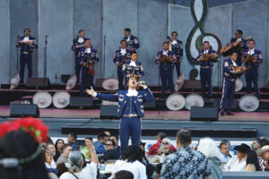 Mariachi band performs on stage with a lead singer reaching toward the crowd; audience sits at tables watching in front of them.