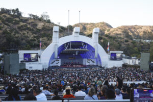Large outdoor amphitheater with a full audience watching a stage performance; surrounding hills and flags visible.