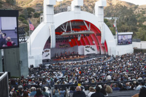 Outdoor amphitheater with a large white stage, red backdrop, and a full crowd watching a live band under a sunny hillside setting.