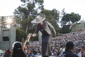 Charro-style performer in a gray suit and oversized sombrero hands a cup to a fan in the front row at a large outdoor stadium concert.