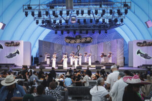 Mariachi band in white outfits performing on a large blue-lit stage, with a crowd watching in the foreground at a festival venue