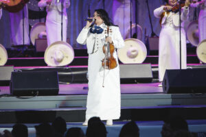 Female mariachi-style singer in a white suit performs on stage, microphone in hand and violin slung over her shoulder, with backup musicians in white in the background.