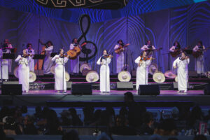 Female mariachi ensemble in white dresses performing on a lit stage with violins and guitars in the background