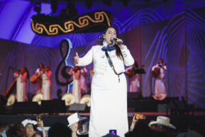 Female singer in a white suit performing on stage with a microphone, backed by musicians under blue lighting.