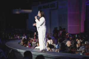 Female singer in a white suit performing on a curved stage, microphone in hand, as a crowd watches in a dimly lit venue with purple lighting.
