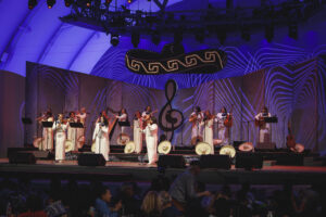 A group of women in white dresses perform music on a blue-lit stage with a large treble-clef backdrop behind them.