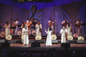 Group of female mariachi musicians on stage in white dresses, guitars and violins, with sombreros arranged at their feet under purple stage lighting.