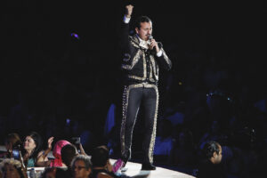 Mariachi-style singer on stage in a black gold-embroidered suit, microphone in hand, fist raised.
