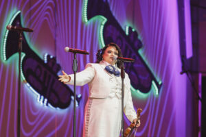Female performer in a white suit sings into a microphone on a colorful stage, holding a violin in one hand.