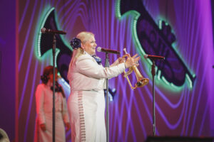 Female trumpeter on stage in a white outfit sings into a microphone while holding a trumpet, with colorful neon backdrop behind.