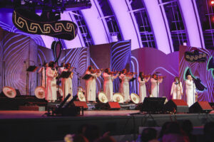 A line of female violinists in white dresses perform on a purple-lit stage with decorative panels in the background.
