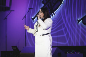 Female singer in a white outfit performs on stage with a microphone, purple lighting in the background.
