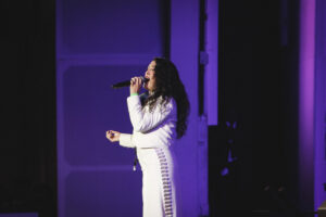 Singer performing on stage, singing into a handheld microphone under purple lights, wearing a white outfit with side stripe details.