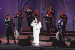 Female singer in a white dress performing center stage with a mariachi band in blue embroidered suits behind her, playing trumpets and guitars, under purple lighting.