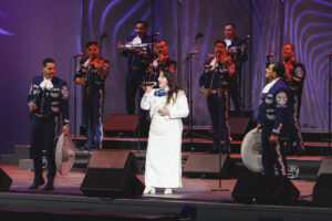 Female singer in a white dress performs on stage with a mariachi band in blue suits, trumpets and guitars visible in the background under purple lighting.