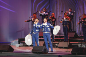 Mariachi band performs on a lit stage; two lead singers in blue embroidered suits hold white sombreros while trumpets and violins play in the background.