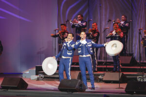 Mariachi band performing on stage; two lead singers in blue outfits with white embroidery sing into microphones, each holding a large white sombrero, with trumpeters in the background on purple-lit stage.