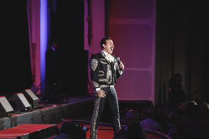 Male singer in an ornate black-and-gold bolero jacket performs on a dimly lit stage with a microphone, purple lighting, and an audience in the foreground.