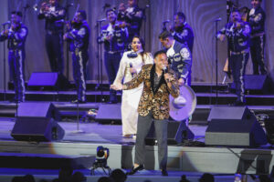 Male singer in a gold patterned jacket performs center stage with a microphone, while a woman in white and a mariachi band stand behind him on a blue-lit stage.