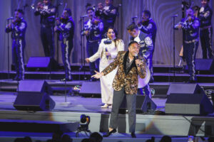 Male singer in a gold floral blazer performs center stage with a female vocalist and a mariachi band behind them.