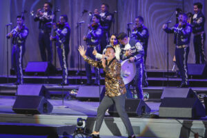 Lead singer in a gold sequin jacket performs center stage with a mariachi band in blue outfits on a lit concert stage.