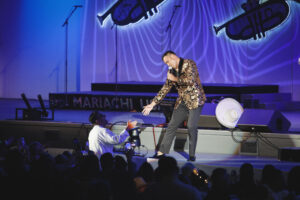 Male singer in a patterned jacket leans toward the crowd on stage, microphone in hand, under blue lights.
