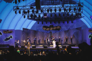 Stage performance with a large band and singers under blue lighting, audience silhouettes in the foreground.