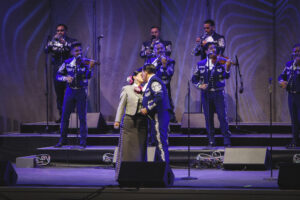Pareja besándose en el centro del escenario mientras una banda de mariachi toca tras ellos.