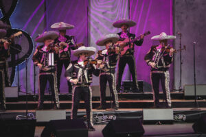 Group of mariachi musicians wearing ornate black suits and large silver sombreros performing on a purple-lit stage.