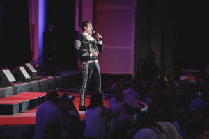 Male mariachi singer in a black and silver embroidered suit on stage with a microphone.psum