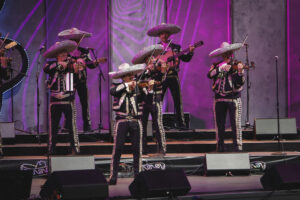 Mariachi band performing on a stage with purple lighting; musicians wear matching black sequined outfits and large sombreros, playing violins and guitar in a live concert.