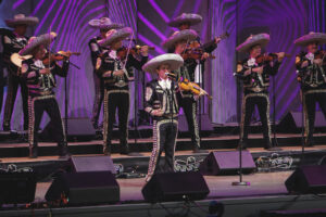Mariachi band on stage in ornate black suits with silver trim and sombreros, performing under purple lights.