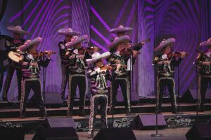 Mariachi musicians in ornate silver-trimmed outfits and large sombreros perform on a stage with purple lighting behind them, playing violins and guitars.
