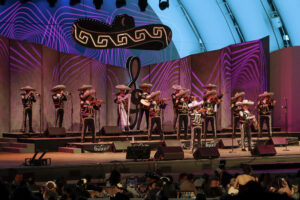 Mariachi band in traditional outfits performing on a colorfully lit stage with a large sombrero prop and neon backdrop, audience in front.