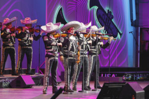 Mariachi band performing on stage in matching black and silver charro outfits with silver sombreros, playing violins under purple stage lights