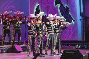 Mariachi musicians performing on a stage in matching black outfits with silver embroidery and white sombreros, playing violins under purple neon lighting.