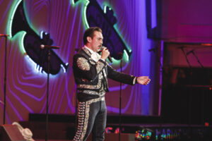 Male mariachi-style singer in an ornate black suit with silver embroidery, singing into a microphone on a neon-lit stage.