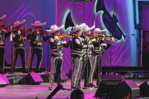Group of mariachi musicians in silver-embellished outfits and white sombreros playing violins on a purple-lit stage.
