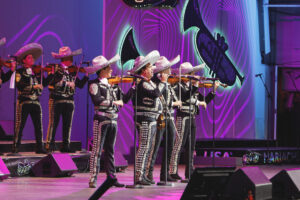 Mariachi band in silver-embellished outfits and sombreros performing on a purple-lit stage with violins, microphones ready at front of stage.