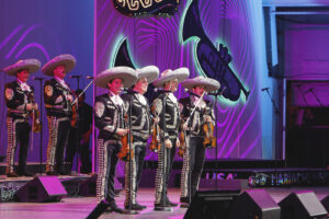 Mariachi band on a stage in matching embroidered charro outfits and sombreros, playing violins with a purple neon backdrop.
