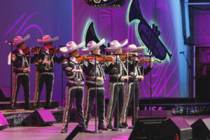 Mariachi band performing on stage, wearing black outfits with silver trim and sombreros, playing violins.