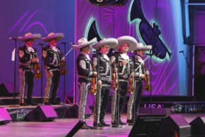 Mariachi band performing on a brightly lit stage in traditional outfits and large sombreros, each musician holding a violin and standing at microphones with purple neon backdrop behind them.