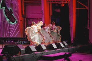 Three dancers in flowing white dresses with orange trim perform a traditional folk dance on a lit stage.