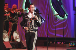Male mariachi singer in a glittering embroidered suit holds a microphone on stage, with violinists in the background and neon lights.