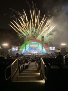Nighttime outdoor concert with a brightly lit, multi-colored stage as fireworks explode overhead and a large crowd watches from stadium seating.