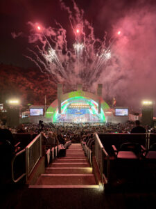 Nighttime outdoor concert with a brightly lit, colorful stage and fireworks exploding above a large audience, stadium seating visible in foreground.